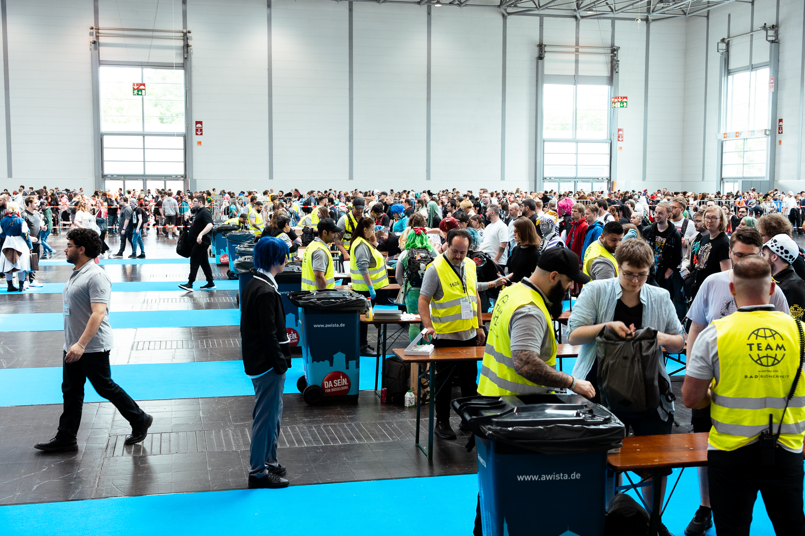 Sicherheitsmitarbeiter in gelben Warnwesten kontrollieren Besucher und Taschen bei einer Großveranstaltung in einer Messehalle.